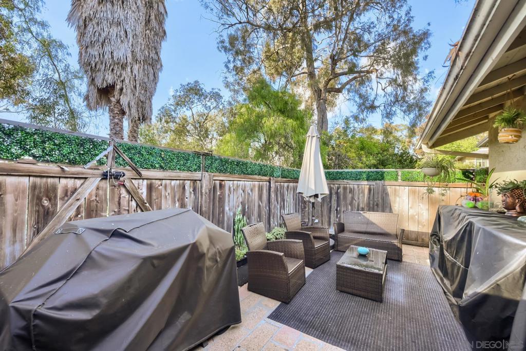 The private patio at 4339 Caminito Del Diamante #60, with wicker lounge seating and an umbrella set on pavers, enclosed by a wood privacy fence beneath a tall eucalyptus canopy in afternoon light